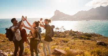 Ready to start hiking? Check out our hiking gear list as well as some resources for finding trails near you before you head out! (Young adult friends on a hike celebrate reaching a summit near the coast, full length, side view)