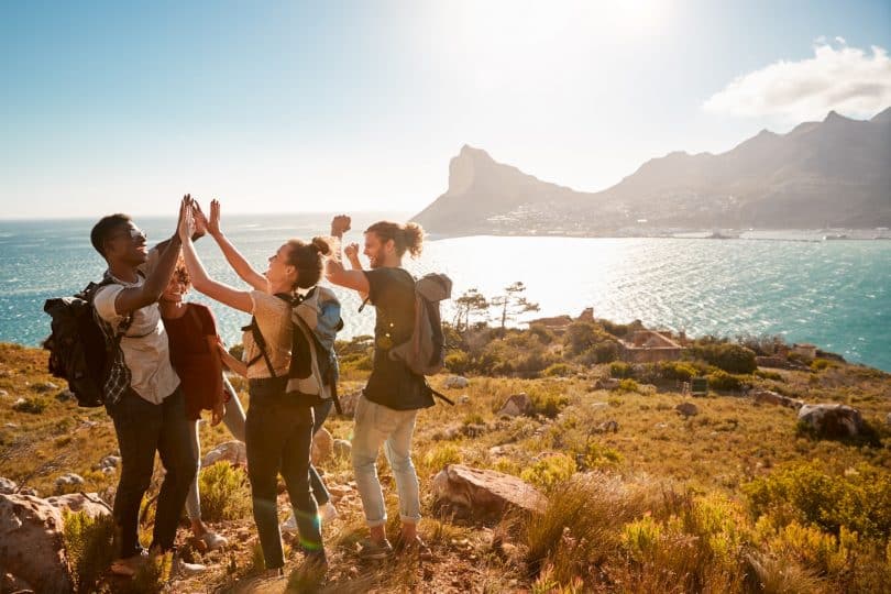 Ready to start hiking? Check out our hiking gear list as well as some resources for finding trails near you before you head out! (Young adult friends on a hike celebrate reaching a summit near the coast, full length, side view)