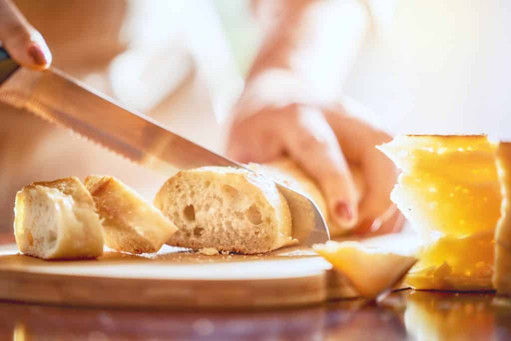 Flour, water, salt, and yeast—it's not quite as easy as it sounds! We'll help you make your first loaf of bread with these bread baking tips for beginners. (Hands of person cutting bread and cheese on wooden table.)