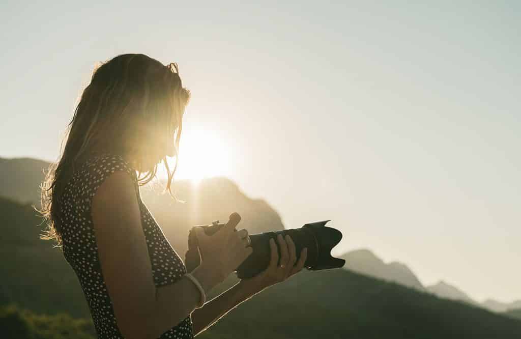 Image shows a woman looking at her DSLR camera with the sunrise in the background.