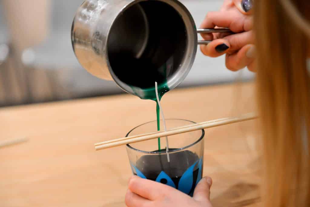 Image shows a girl making a candle, pouring blue wax into a glass, with a wick held in the center by two wooden sticks.